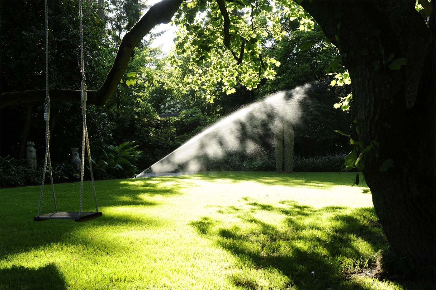 Sprinkleranlage, die Rasen im Garten bewässert, mit Schatten von Bäumen im Hintergrund.