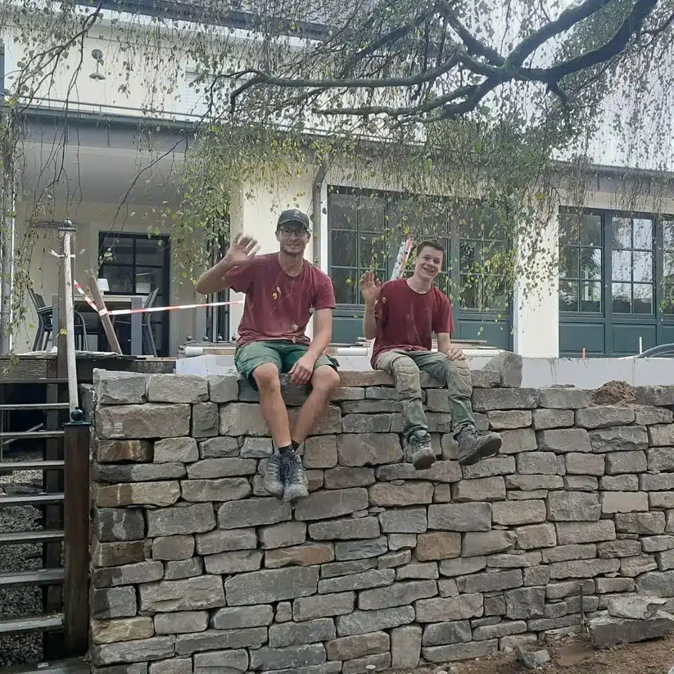 Gartengestaltung Kleve, 2 Kollegen auf Steinmauer, rot, grün Zwei Kollegen stehen auf einer Steinmauer in Kleve, tragen rote Arbeitskleidung und winken mit grünem Umfeld im Hintergrund.