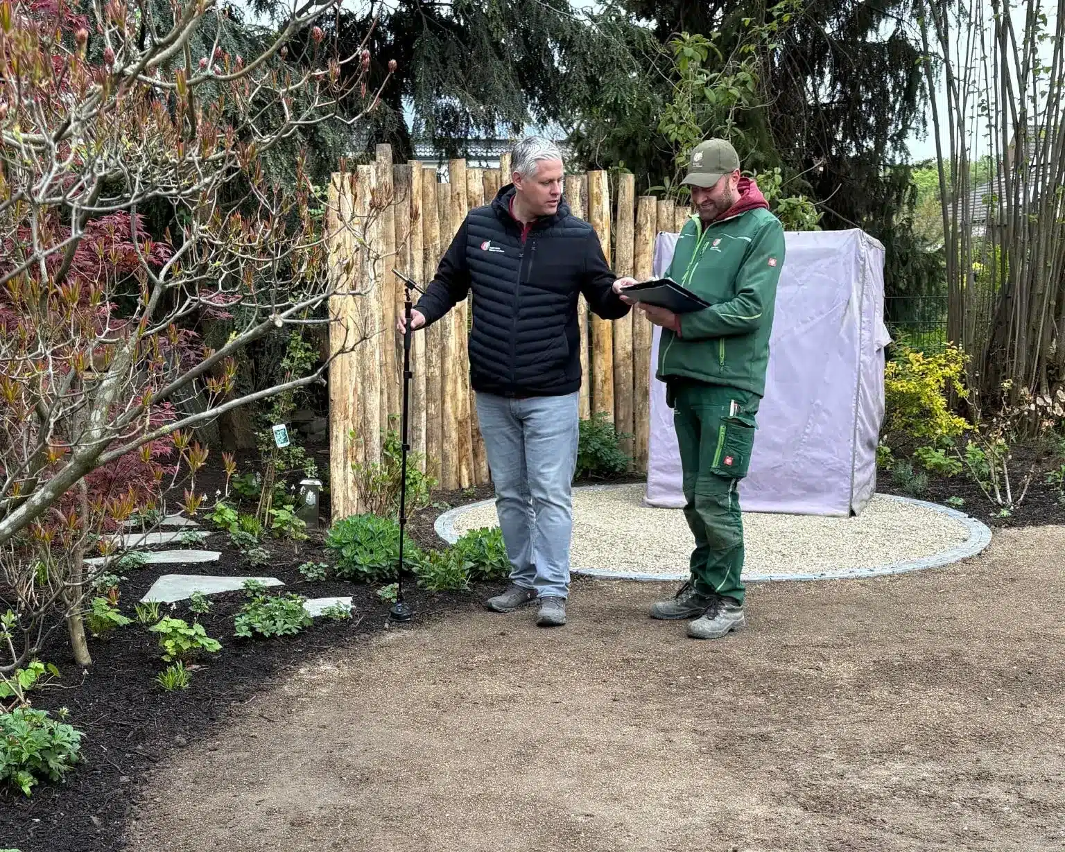 Kleve Gartengespräch, Baustellenbesprechung im Garten mit grünen und schwarzen Arbeitsoutfits Vier Frauen in roten Poloshirts posieren auf einer Steinmauer in einem Garten mit Pflanzen im Hintergrund.