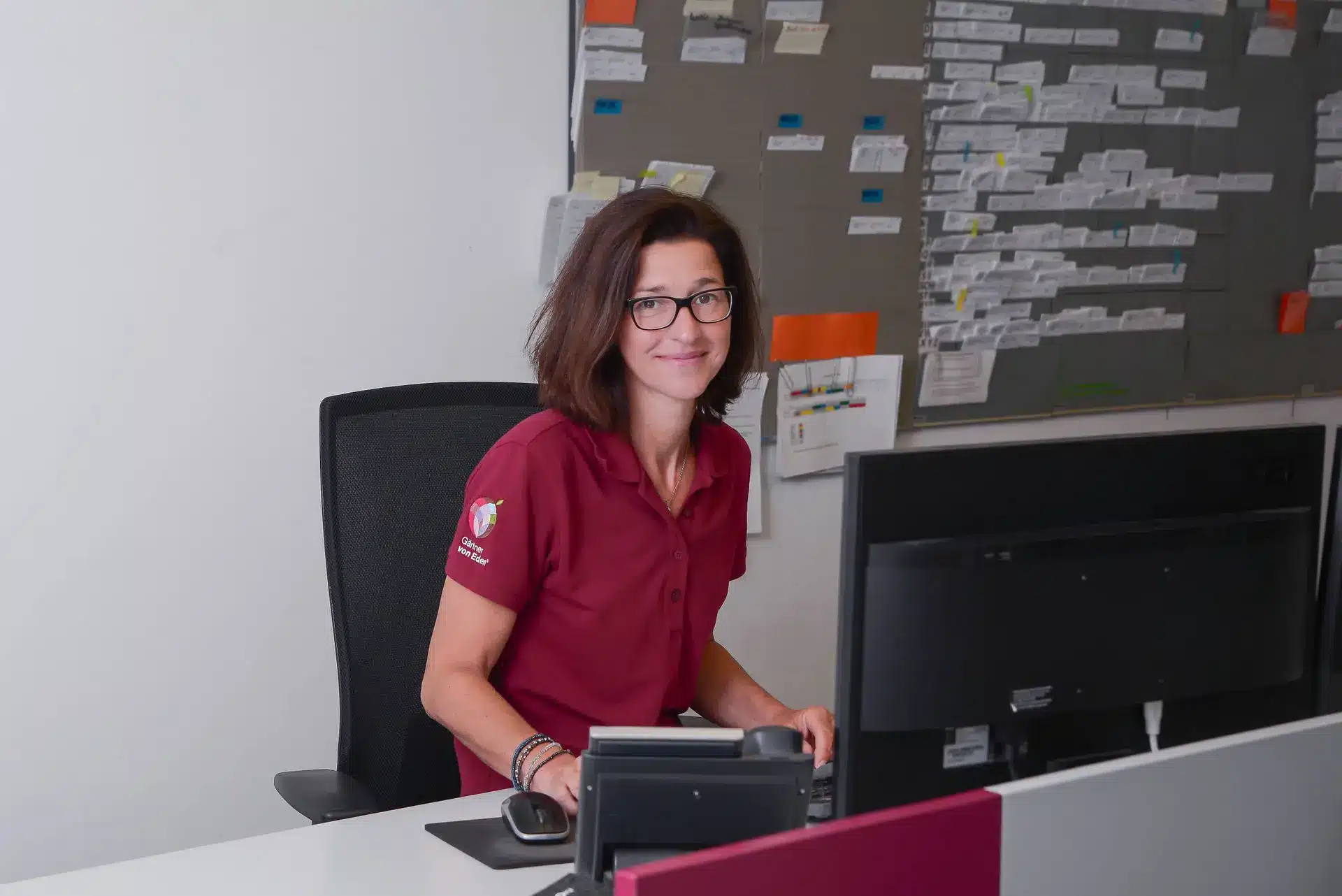 Gartengestaltung Kleve, Mitarbeiterin Büro, rote Arbeitskleidung, Brille In einem Büro sitzt eine Mitarbeiterin in roter Arbeitskleidung mit Brille vor einem Monitor und blickt freundlich in die Kamera.