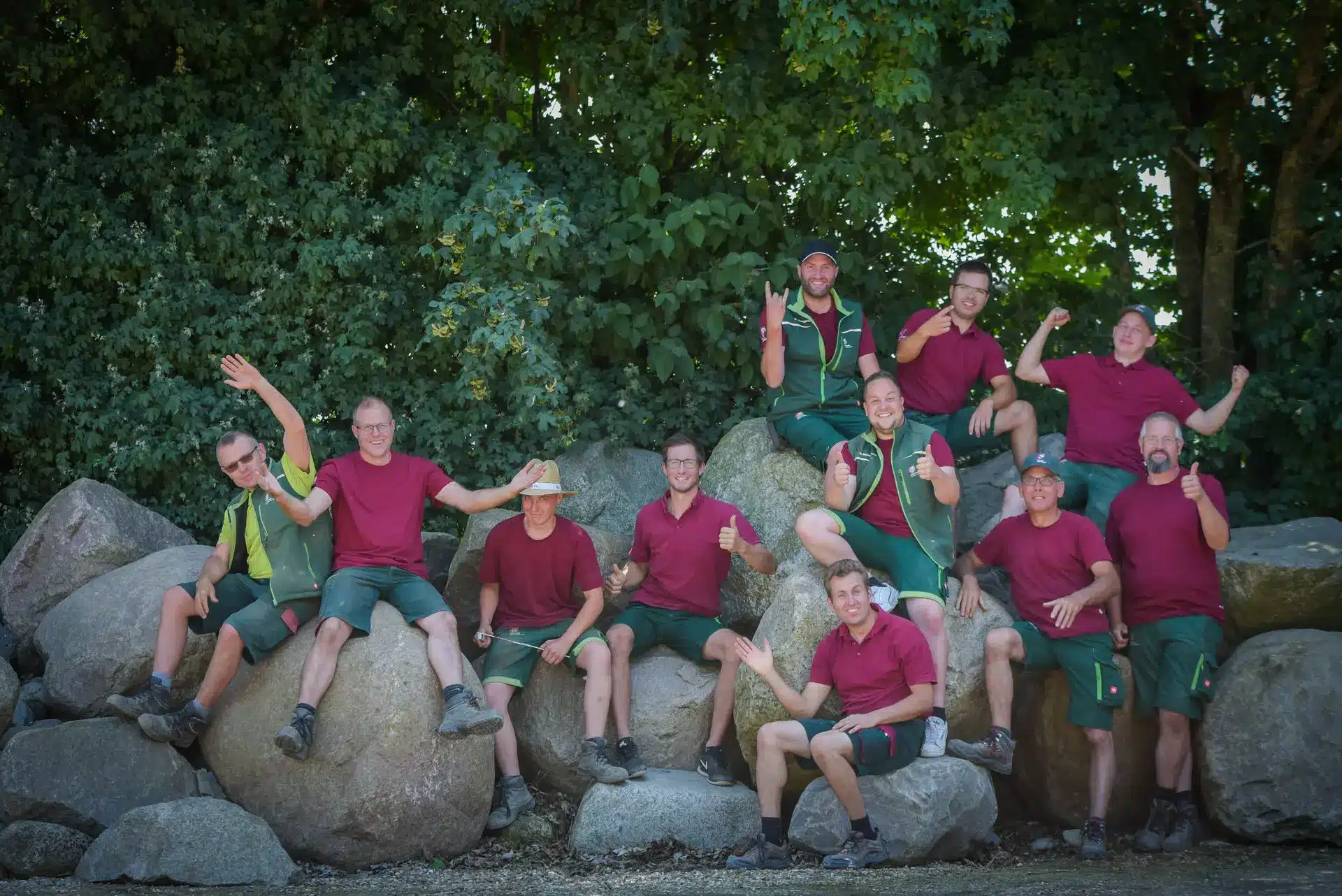 Gartengestaltung Kleve Teamfoto, Steingruppe, grüne Arbeitskleidung, Outdoor Mehrere Mitarbeitende in grünen Arbeitslatzhosen und T-Shirts posieren gemeinsam auf einer Steingruppe vor dichtem Grün in Kleve.
