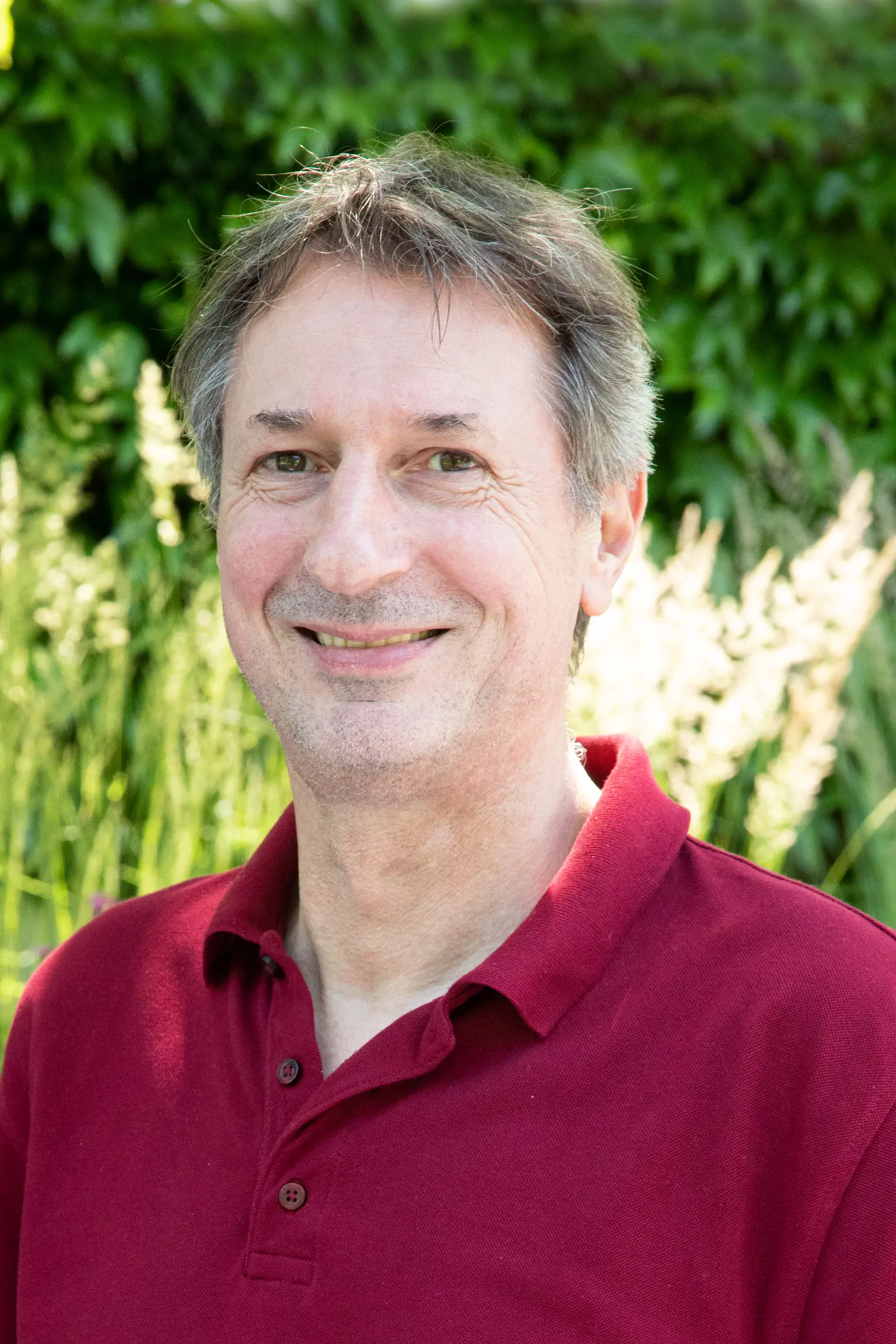 Mann mit braunen Haaren in einem roten Poloshirt steht vor grünen Pflanzen im Garten.