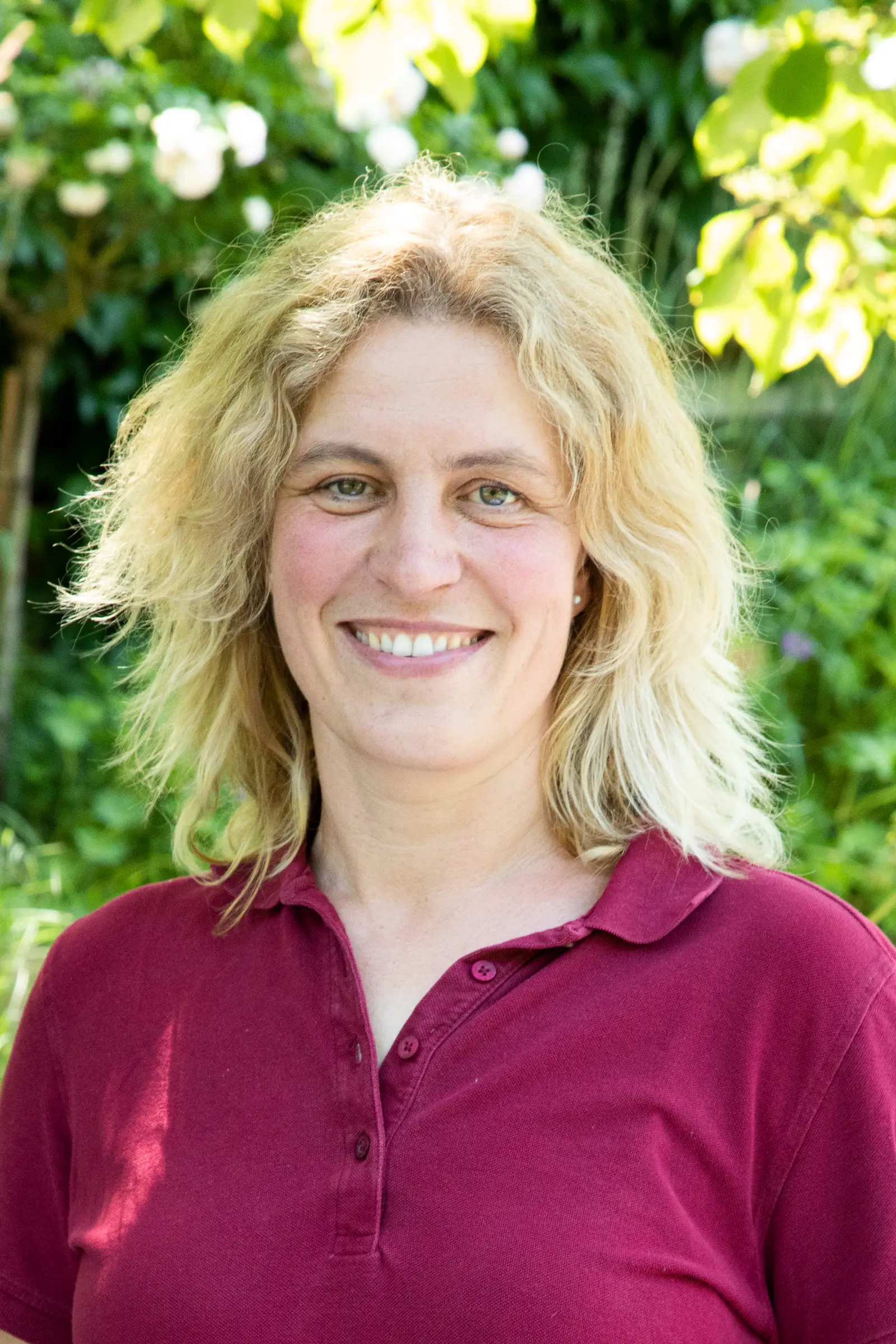 Lächelnde Frau mit blonden, lockigen Haaren in einem roten Poloshirt vor grüner Vegetation.