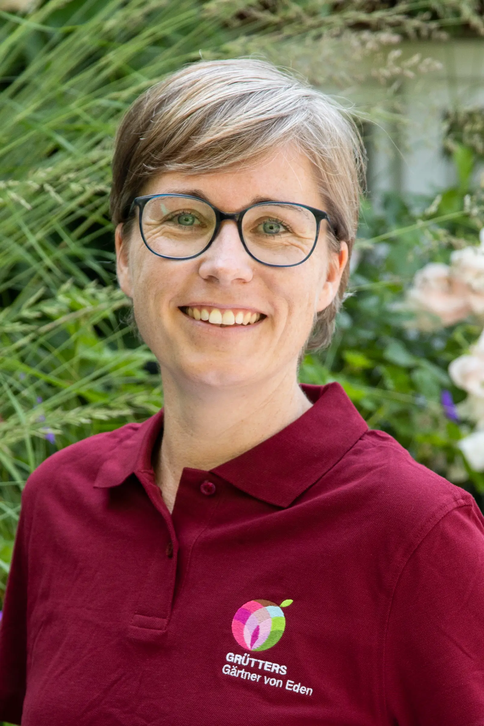 Frau mit kurzen blonden Haaren und Brille trägt ein rotes Poloshirt mit einem Logo.