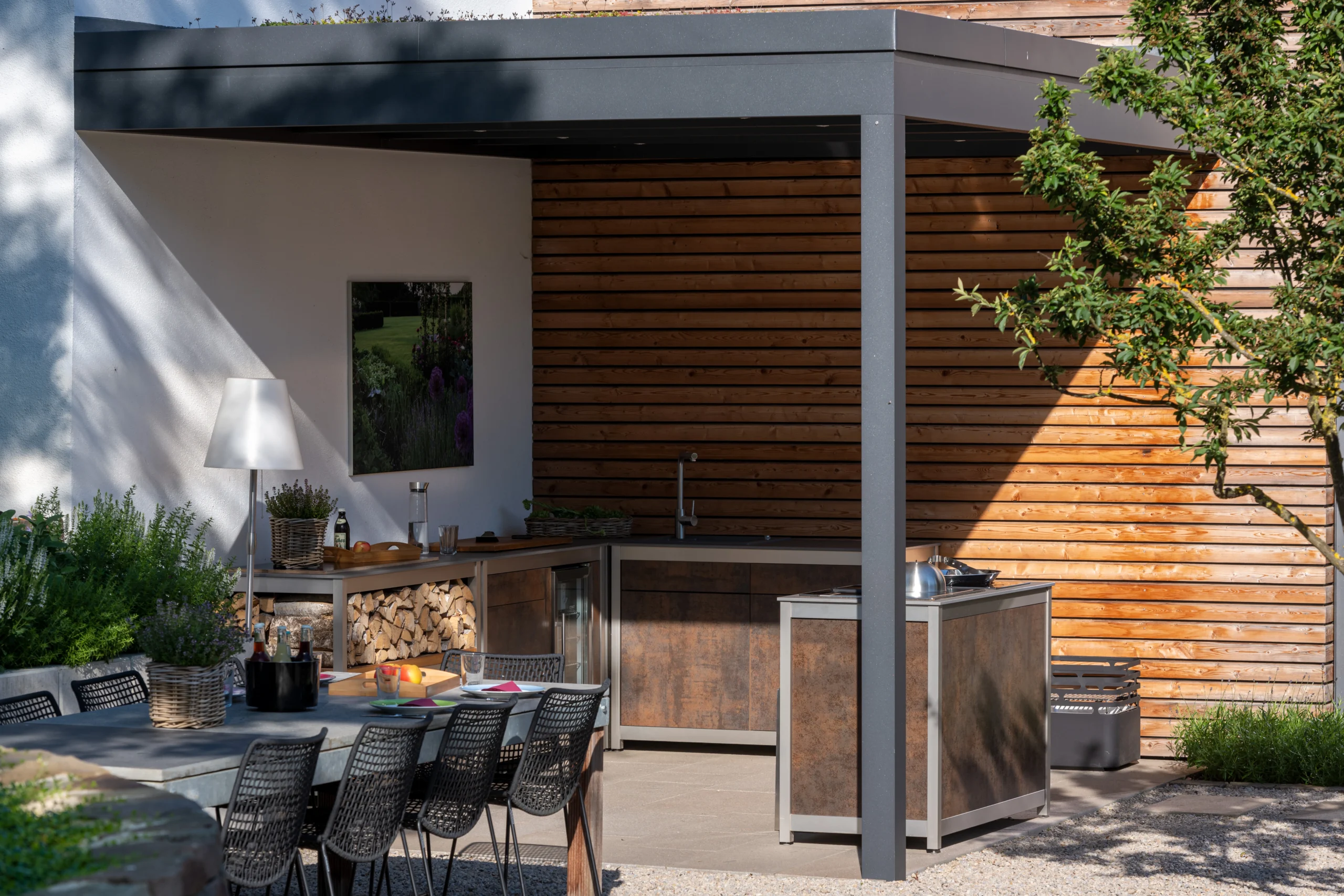 Überdachte Außenküche mit Holzverkleidung und Garten-Essbereich Unter einer modernen Überdachung steht eine Außenküche mit Arbeitsflächen, Spüle und angrenzendem Esstisch im Garten.