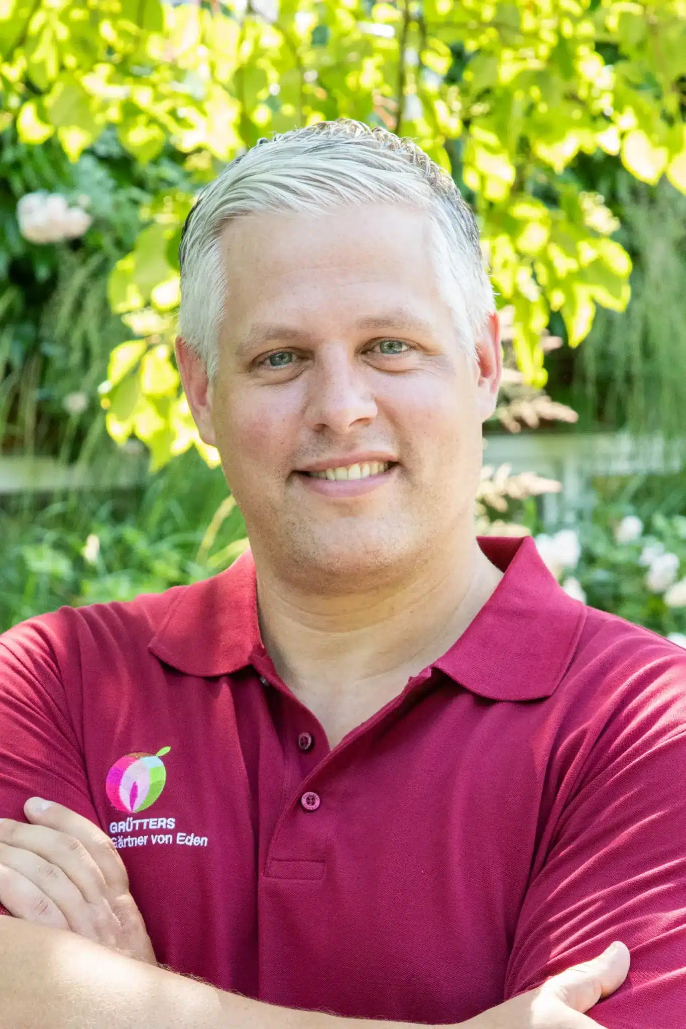 Lächelnde Frau mit blonden, lockigen Haaren in einem roten Poloshirt vor grüner Vegetation.