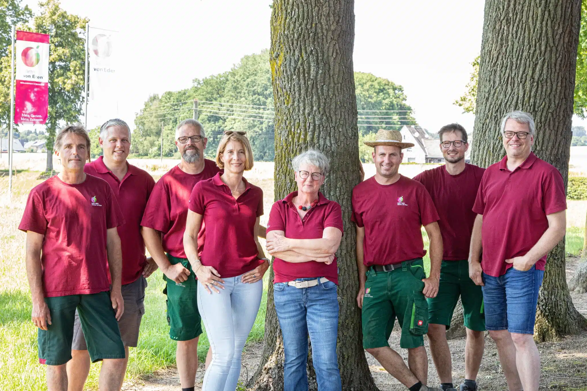 Eine Gruppe von acht Personen steht vor einem Unternehmensschild in einem Gartenbereich.