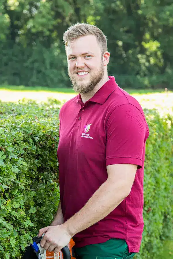 Ein Mann mit Brille und Kappe lächelt in die Kamera, während er ein rotes T-Shirt trägt.