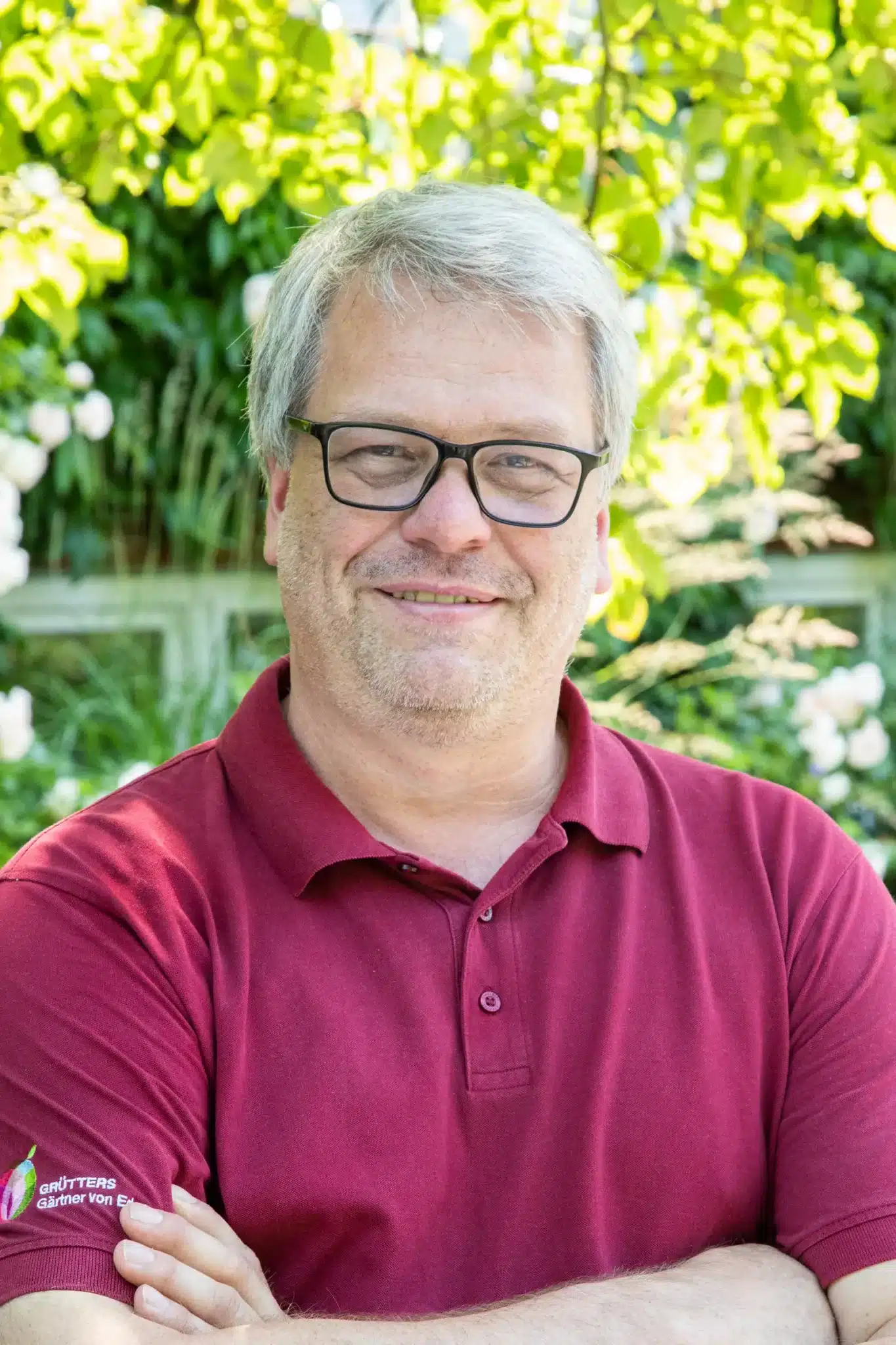 Mann mit grauen Haaren in einem roten Poloshirt steht vor grüner Vegetation.