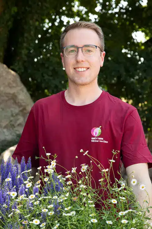 Ein Mann mit Brille und Kappe lächelt in die Kamera, während er ein rotes T-Shirt trägt.