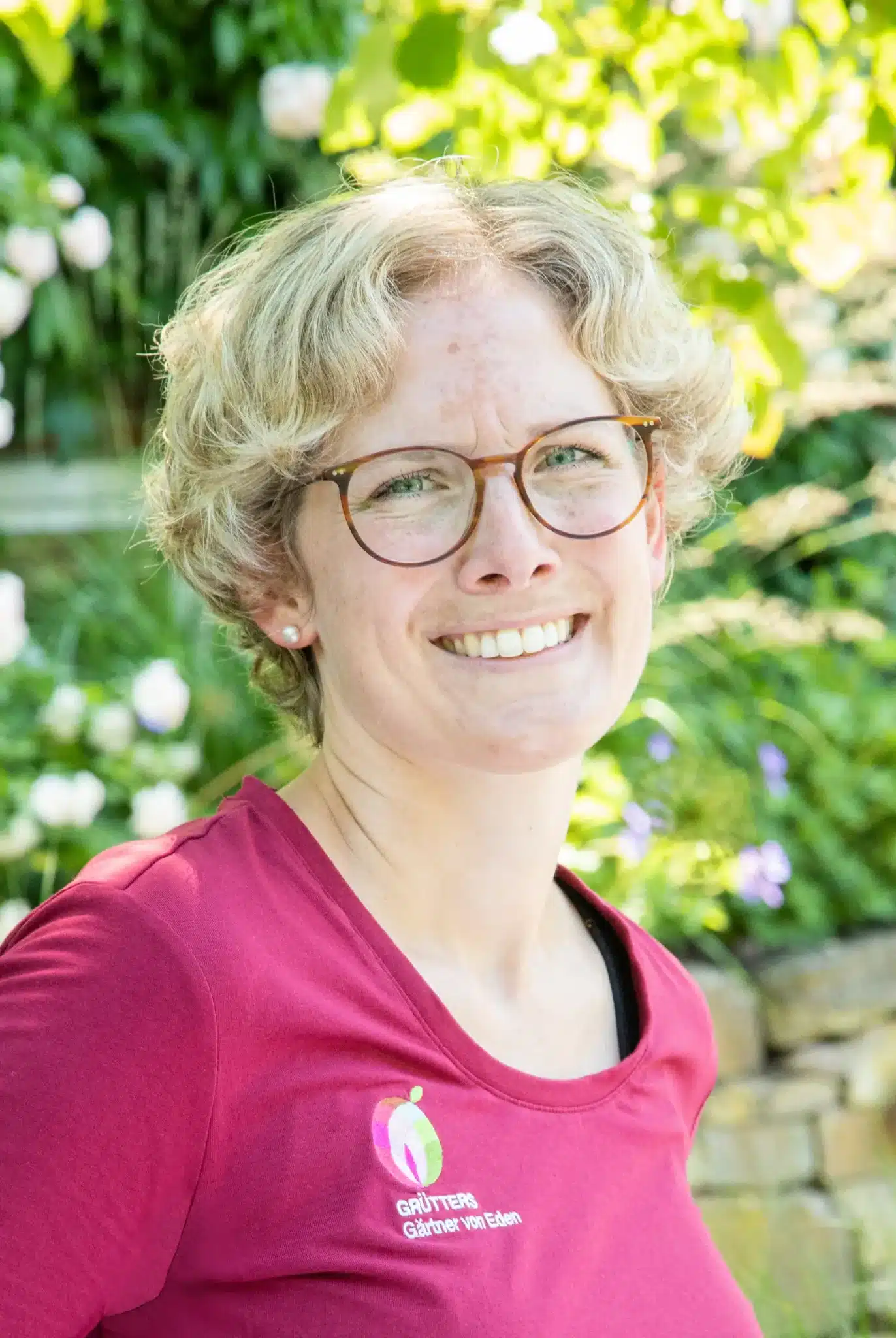 Frau mit blonden, lockigen Haaren in einem roten Poloshirt vor grüner Vegetation.