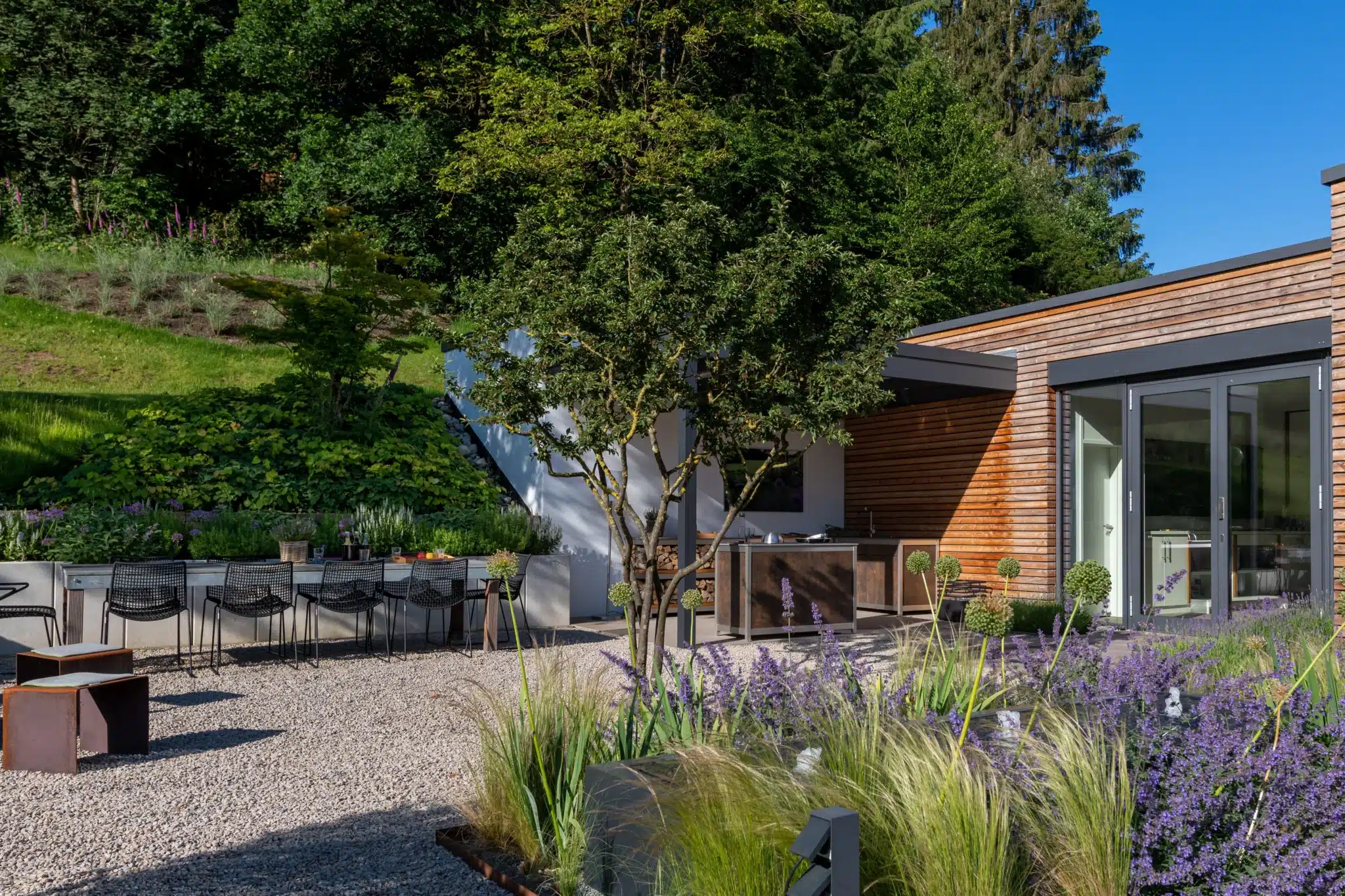 Außenküche im Garten mit Essplatz auf der Kiesterrasse Außenküche vor einer Holzfassade mit großem Essplatz auf einer Kiesterrasse, umgeben von Gräsern und violett blühenden Stauden.